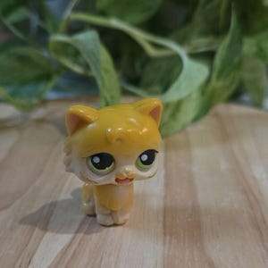 May include: A small, yellow cat figurine with a light tan face and paws. The cat has large, expressive green eyes and a small pink tongue. The figurine is standing on a wooden surface, with green foliage in the background.
