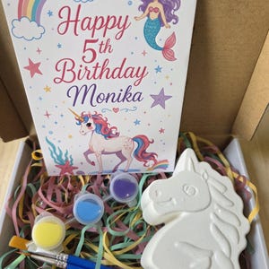 May include: A unicorn-themed craft kit in a cardboard box. The kit includes a white unicorn figure, paint pots in yellow, blue, and purple, paintbrushes, and a birthday card with a rainbow, mermaid, and unicorn. The card reads "Happy 5th Birthday Monika."