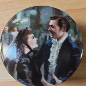 May include: A round decorative plate featuring a romantic scene with two people in period clothing. The man wears a dark suit with a white ruffled shirt, and the woman wears a black dress. The plate has a glossy finish.