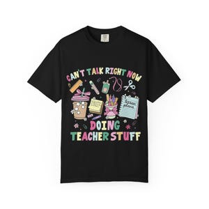 May include: A black short-sleeved t-shirt featuring a colourful graphic print with the text "CAN'T TALK RIGHT NOW DOING TEACHER STUFF". The design includes illustrations of a coffee cup, a notepad, a pencil holder, scissors, and a ruler.