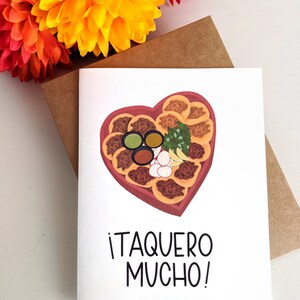 May include: A white card with a heart-shaped illustration of a taco platter with toppings. The text on the card reads "&iexcl;Taquero Mucho!"