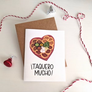May include: A white card with a red heart-shaped container filled with tacos, salsa, onions, and cilantro. The text on the card reads "&iexcl;Taquero Mucho!"