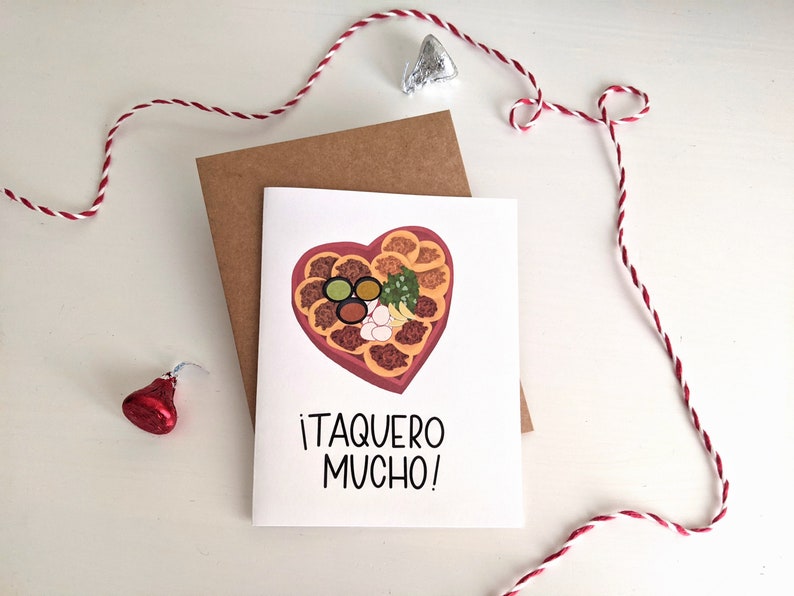May include: A white card with a red heart-shaped container filled with tacos, salsa, onions, and cilantro. The text on the card reads "&iexcl;Taquero Mucho!"