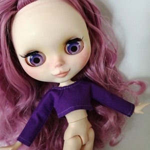 May include: A doll with long, curly, pink hair wearing a purple crop top with long sleeves.