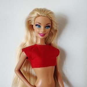 May include: A blonde doll wearing a red crop top and a nude-colored bottom. The doll has blue eyes and pink lips.