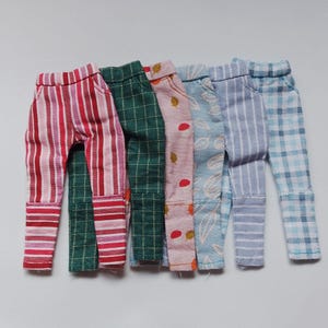May include: Five pairs of doll-sized pants in various patterns, including red and white stripes, green plaid, pink floral, blue stripes, and blue plaid.