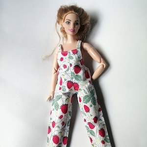 May include: A doll wearing a white and green jumpsuit with a strawberry print. The jumpsuit has thin straps and a wide leg.