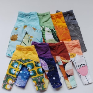 May include: A collection of eight pairs of colourful patterned trousers for dolls. The trousers are made from different fabrics and have various designs, including floral, geometric, and abstract patterns.