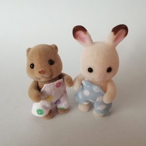 Op de afbeelding: Twee pluche speelgoeddieren die pyjama's dragen. Een bruine beer met een witte pyjama met veelkleurige stippen en een wit konijn met een blauwe pyjama met witte stippen.