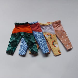 May include: Four pairs of colorful patterned doll pants. The pants are made of different fabrics and have a variety of designs, including geometric patterns, floral prints, and polka dots.
