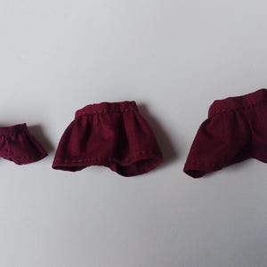 May include: Three burgundy doll skirts of different sizes. The skirts are made of a soft, textured fabric and have a simple, gathered design.