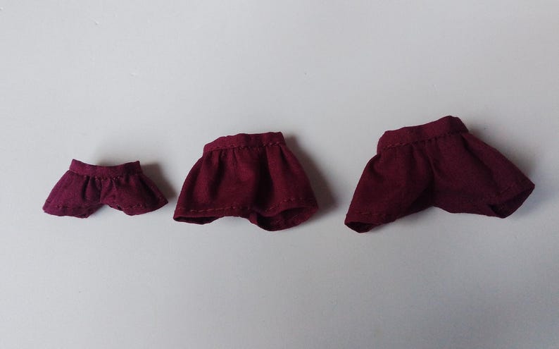 May include: Three burgundy doll skirts of different sizes. The skirts are made of a soft, textured fabric and have a simple, gathered design.
