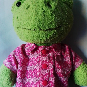 May include: A green plush frog wearing a pink and white patterned shirt with red buttons.