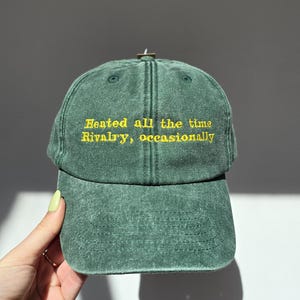 May include: A green baseball cap with the embroidered text "Heated all the time, Rivalry, occasionally" in yellow. The cap is made of a textured fabric and has a curved brim.