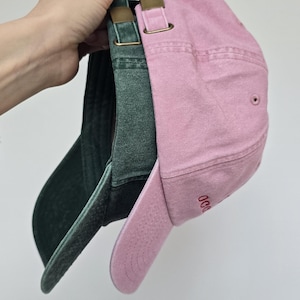 May include: Three baseball caps in shades of pink and green are displayed. The caps have curved visors and adjustable straps with brass-colored buckles. The pink cap has embroidered text on the side. The caps are made of a soft, textured fabric.