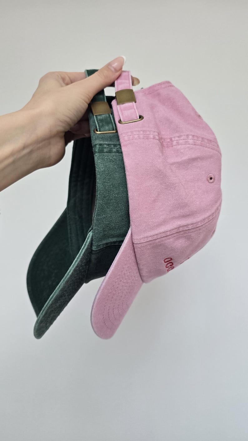 May include: Three baseball caps in shades of pink and green are displayed. The caps have curved visors and adjustable straps with brass-colored buckles. The pink cap has embroidered text on the side. The caps are made of a soft, textured fabric.