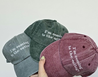 May include: Three baseball caps in washed-out shades of gray, dark green, and maroon. Each cap features white embroidered text that reads, I'm coming to the cottage, positioned on the front panel above the curved brim with visible stitching.