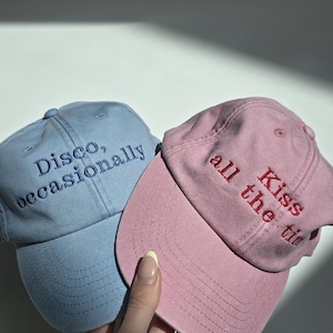 May include: Two baseball caps, one light blue and one pink, are displayed. The light blue cap has the embroidered text "Disco, occasionally" in blue. The pink cap has the embroidered text "Kiss all the time" in red. The caps are made of a soft fabric.