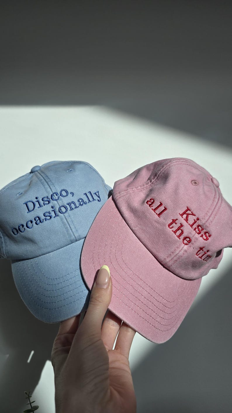 May include: Two baseball caps, one light blue and one pink, are displayed. The light blue cap has the embroidered text "Disco, occasionally" in blue. The pink cap has the embroidered text "Kiss all the time" in red. The caps are made of a soft fabric.