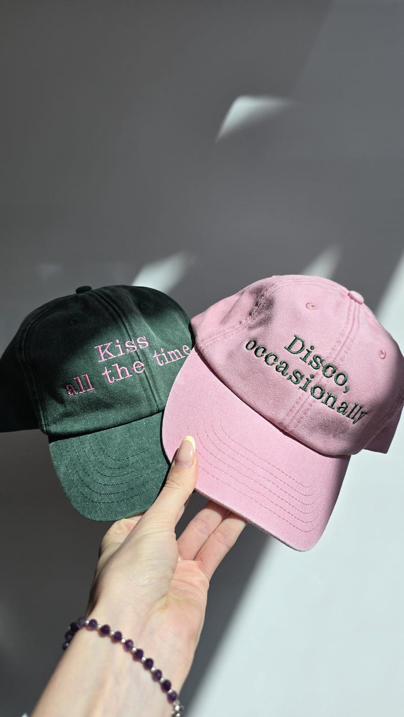 May include: Two baseball caps, one dark green and one pink, are displayed. The dark green cap has the text "Kiss all the time" embroidered in pink. The pink cap has the text "Disco, occasionally" embroidered in green.