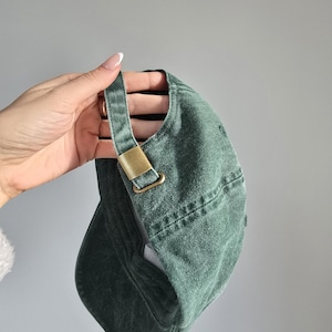 May include: A green baseball cap with a curved brim and adjustable strap. The cap features a brass-colored metal buckle on the strap. The cap is made of a textured fabric.