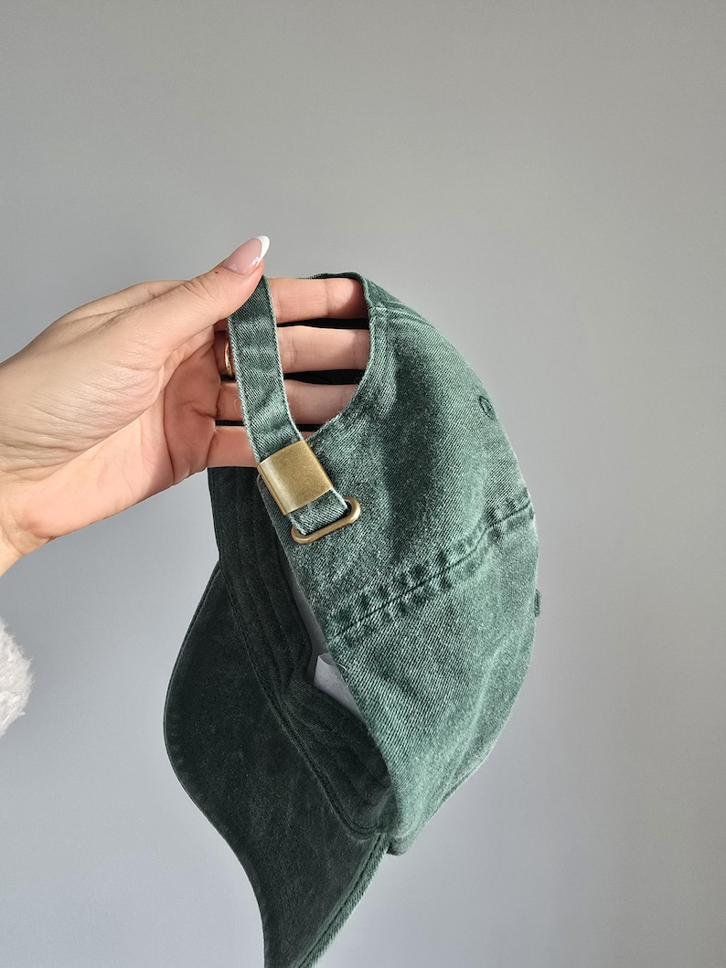 May include: A green baseball cap with a curved brim and adjustable strap. The cap features a brass-colored metal buckle on the strap. The cap is made of a textured fabric.