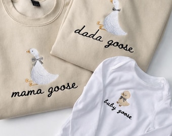 May include: Three matching family outfits: two beige sweatshirts and a white babygrow. Each item has an embroidered goose design with a bow tie. The sweatshirts read "mama goose" and "dada goose", while the babygrow says "baby goose".