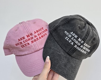 May include: Two baseball caps, one dusty rose and one charcoal gray, are held up against a white background. Both caps have the embroidered text "ASK ME ABOUT OUR LORD & SAVIOR ILYA ROZANOV" in contrasting colors.