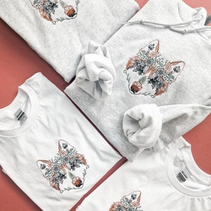 Puede incluir: Tres camisas con un diseño de lobo floral. Dos son camisetas blancas y una es una sudadera con capucha gris.