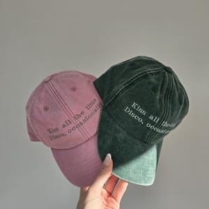 Puede incluir: Dos gorras de béisbol, una rosa empolvado y otra verde oscuro, se muestran. Las gorras tienen viseras curvas y el texto "Kiss all the time, Disco, occasionally" está impreso.