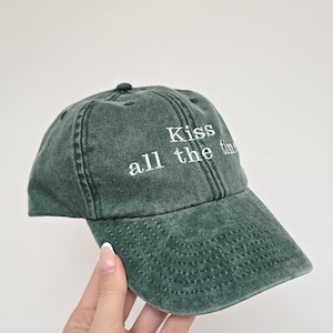 May include: A dark green baseball cap with the embroidered text "Kiss all the time" in white. The cap has a curved brim and a classic six-panel design. The hat is made of a soft, textured fabric.