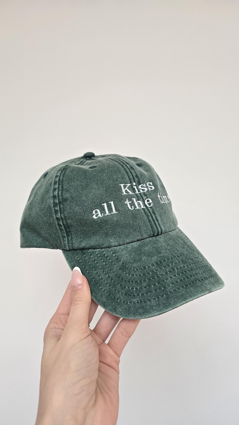 May include: A dark green baseball cap with the embroidered text "Kiss all the time" in white. The cap has a curved brim and a classic six-panel design. The hat is made of a soft, textured fabric.