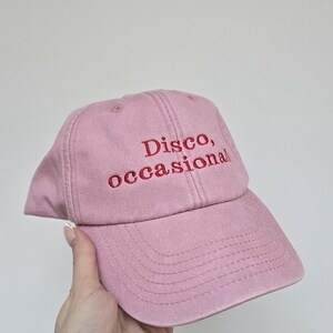 May include: A dusty rose baseball cap with the embroidered text "Disco, occasional" in red thread. The cap has a curved brim and a classic six-panel design, suitable for casual wear.
