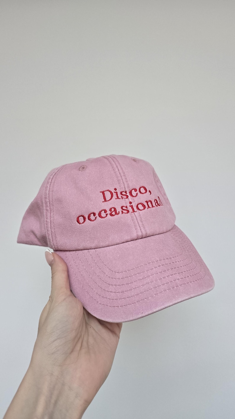 May include: A dusty rose baseball cap with the embroidered text "Disco, occasional" in red thread. The cap has a curved brim and a classic six-panel design, suitable for casual wear.