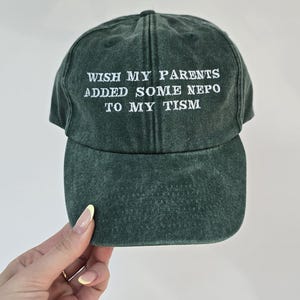 May include: A dark green baseball cap with white embroidered text. The text reads "WISH MY PARENTS ADDED SOME NEPO TO MY TISM". The cap has a curved peak and a classic six-panel design.