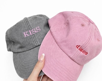 May include: Two baseball caps, one gray and one pink, are held up against a white background. The gray cap has the word "KISS" embroidered in pink. The pink cap has the word "disco" embroidered in red.