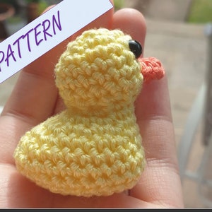 May include: A crocheted yellow duck with a black eye and an orange beak. The duck is sitting on a hand.