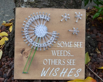 May include: A wooden sign with a white dandelion design made with string wrapped around nails. The sign also has the text "Some see weeds... Others see wishes" in white lettering.