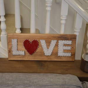 Puede incluir: Un letrero de madera con la palabra "LOVE" escrita en hilo blanco envuelto alrededor de clavos. Un corazón rojo está en el centro del letrero.