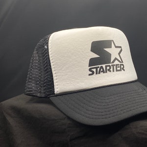 May include: A black and white trucker hat with a white mesh back. The front of the hat features a black and white logo with the word "STARTER" in a bold font.