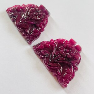 Puede incluir: Dos piedras preciosas de rubí talladas con un diseño floral. Las piedras preciosas son de color rojo intenso y tienen forma triangular.