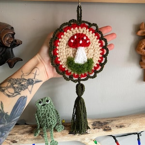 May include: A circular crochet wall hanging featuring a central red and white mushroom motif on a beige background, surrounded by a red and dark green scalloped border. The piece includes a dark green yarn tassel hanging from the bottom.