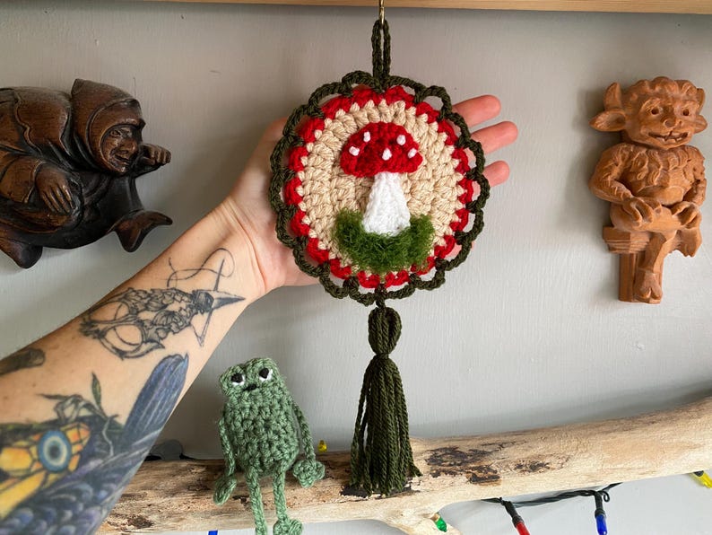May include: A circular crochet wall hanging featuring a central red and white mushroom motif on a beige background, surrounded by a red and dark green scalloped border. The piece includes a dark green yarn tassel hanging from the bottom.