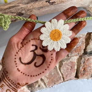 May include: A crocheted daisy with a yellow center and white petals is held in a hand with henna designs. A green crocheted vine with leaves and a yellow measuring tape are also visible. The hand has a henna tattoo of the Om symbol.