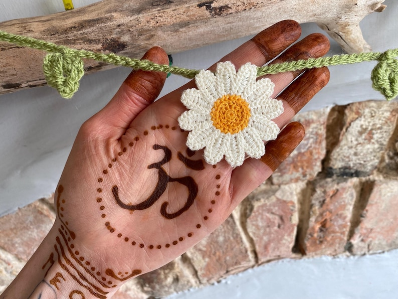 May include: A crocheted daisy with a yellow center and white petals is held in a hand with henna designs. A green crocheted vine with leaves and a yellow measuring tape are also visible. The hand has a henna tattoo of the Om symbol.