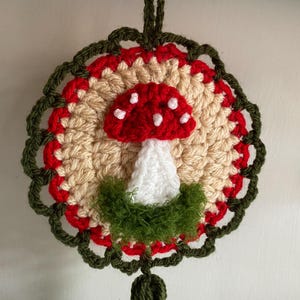 May include: A circular crochet wall hanging featuring a central red and white mushroom motif with white spots. The mushroom is surrounded by a beige circular base, a red border, and an outer dark green scalloped edge with fuzzy green yarn.