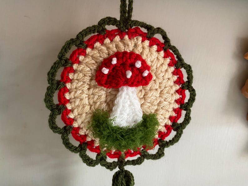 May include: A circular crochet wall hanging featuring a central red and white mushroom motif with white spots. The mushroom is surrounded by a beige circular base, a red border, and an outer dark green scalloped edge with fuzzy green yarn.