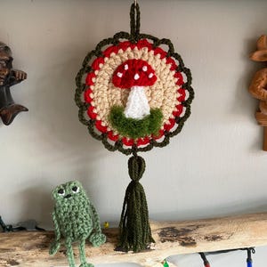 May include: A circular crochet wall hanging featuring a red and white mushroom motif on a beige background, framed by a scalloped dark green border. The piece includes a dark green yarn tassel at the bottom and a loop at the top for hanging.