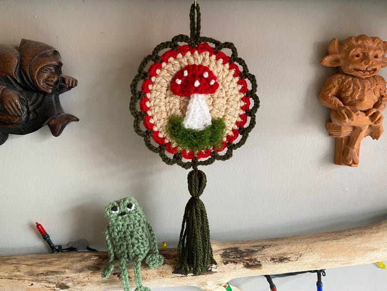 May include: A circular crochet wall hanging featuring a red and white mushroom motif on a beige background, framed by a scalloped dark green border. The piece includes a dark green yarn tassel at the bottom and a loop at the top for hanging.