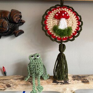 May include: A round crochet wall hanging featuring a red and white mushroom motif with a green base, surrounded by a red and dark green scalloped border and a long dark green tassel. Below it sits a small, light green crocheted frog figurine.